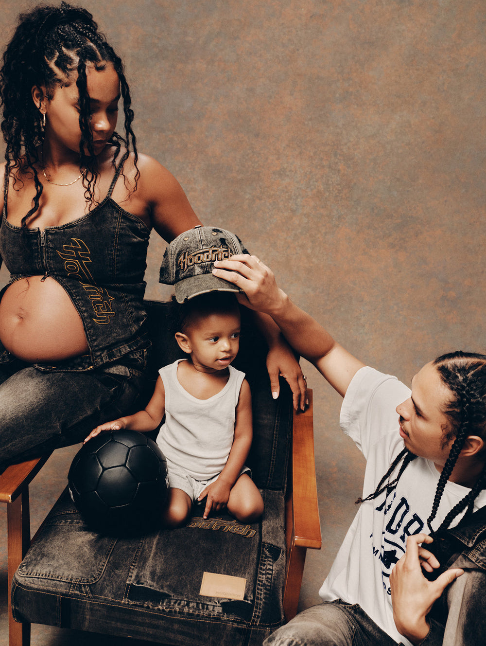 A pregnant woman in a denim 'HOODRICH' corset and matching jeans sits on a wooden chair next to a toddler in a white tank top, while a man adjusts the toddler's baseball cap. A black soccer ball rests on the chair, set against a textured brown studio backdrop.