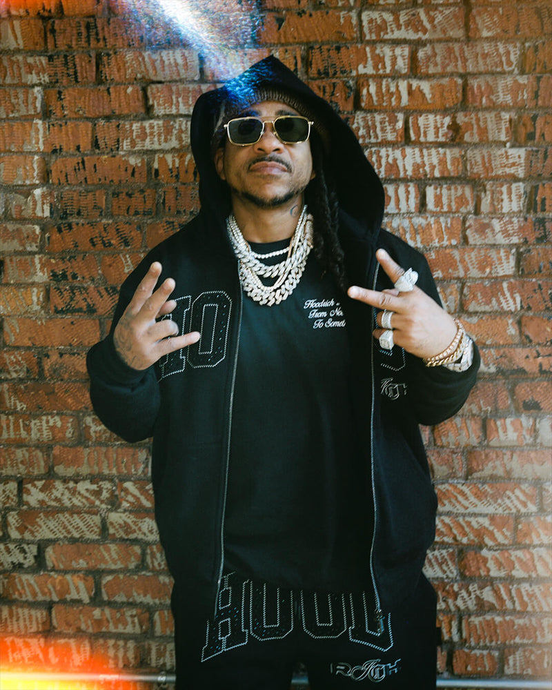 A man wearing a black hoodie, black t-shirt, and black pants stands in front of a brick wall, making hand gestures. He is wearing gold-framed sunglasses, layered diamanté necklaces, and rings.