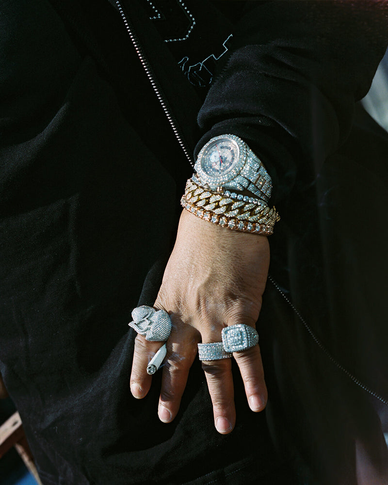 A close-up of a person's hand wearing multiple heavy jewelry pieces, including a large watch, a gold chain bracelet, a silver-toned chain bracelet, three statement rings, and a small pendant, while wearing a black zip-up jacket.