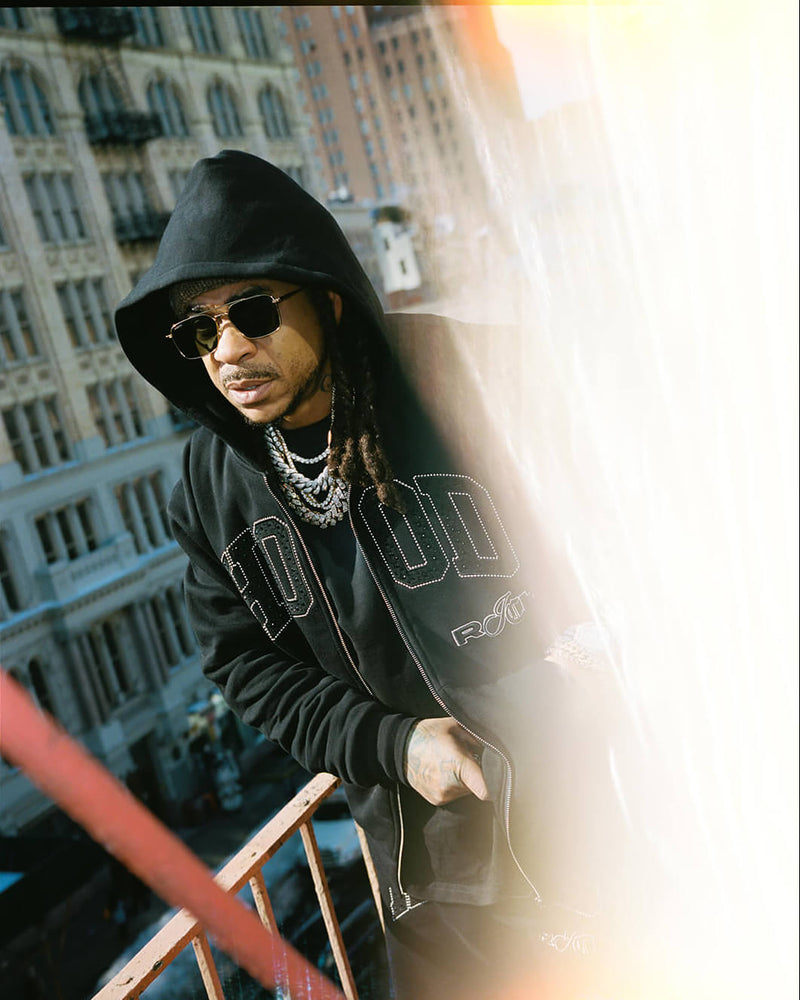 A man wearing a black hooded zip-up sweatshirt with white outlined lettering, black sunglasses, and layered silver chains, standing on an outdoor balcony with city buildings in the background and a strong bright light flare on the right side of the frame.