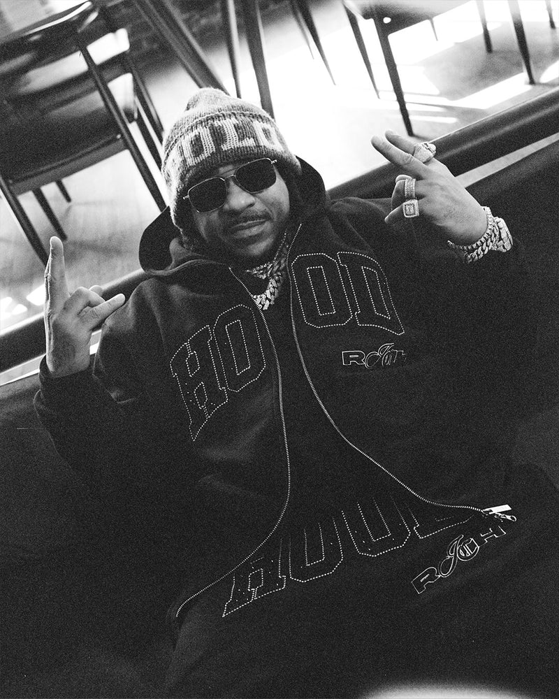 A black and white lifestyle photograph of a man wearing a black hoodie with 'HOOD' embroidered on the front, a knit beanie, and sunglasses, posing with hand gestures.