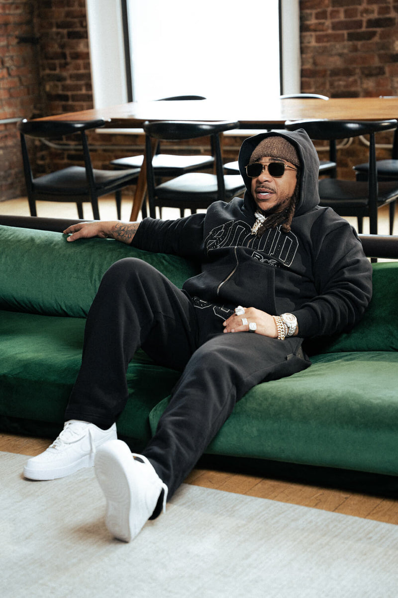 Person sitting on a green couch wearing a black hoodie and sunglasses in a casual setting.